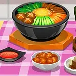 En bordsetting med forskjellige asiatiske retter, inkludert en stor bolle med grønnsaker, en tallerken med kimchi, en kopp grønn te, og flere småretter på en rutete duk