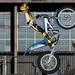 Motorsyklist i gult og svart antrekk balanserer på bakhjulet av en motorsykkel foran et industribygg